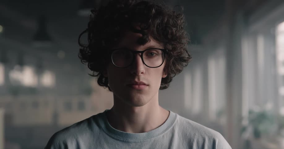 A teenage boy with curly dark hair and glasses looks ahead