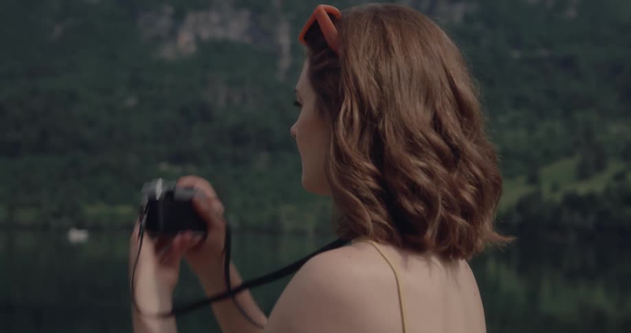 A woman is standing near shallow water in a forest and lifts her camera to her face to take a picture