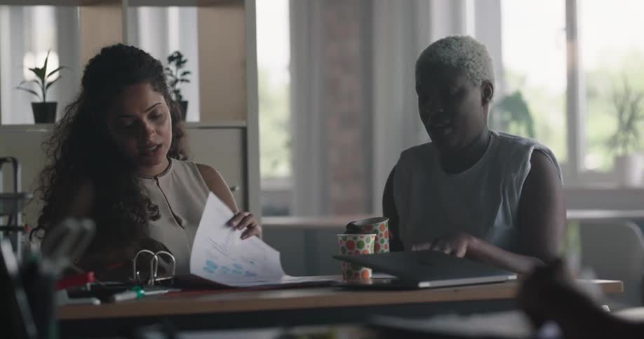 Two women sitting in an office space discussing work smiling and having a snack