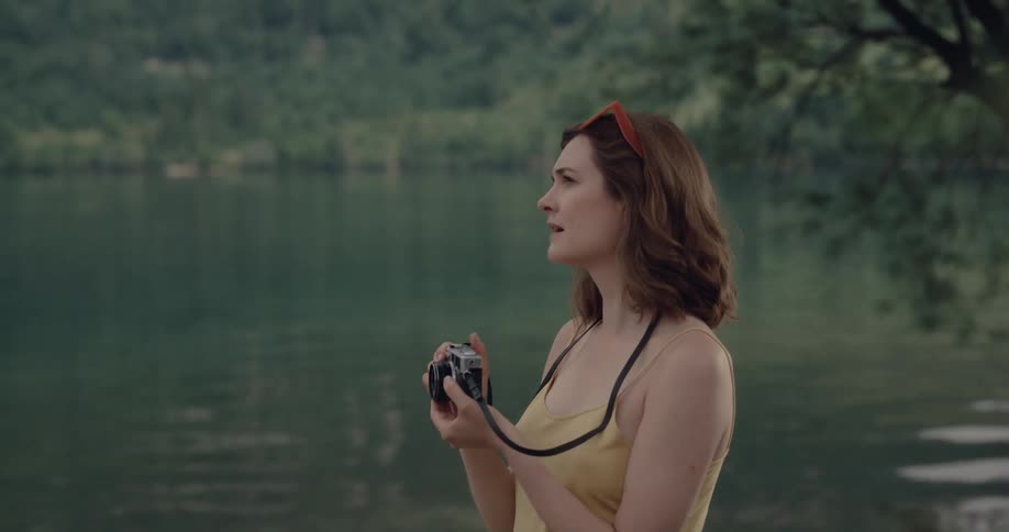 Woman with wavy brown hair yellow shirt and green pants stands by lake and photographs water and mountain with forest