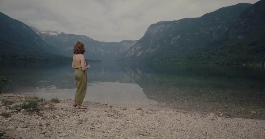 Woman with wavy brown hair yellow shirt and green pants walks by lake and photographs water and mountain with forest