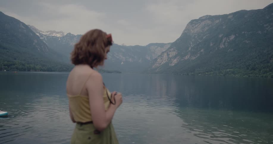 Woman with wavy brown hair yellow shirt and green pants stands by lake and photographs water and mountains