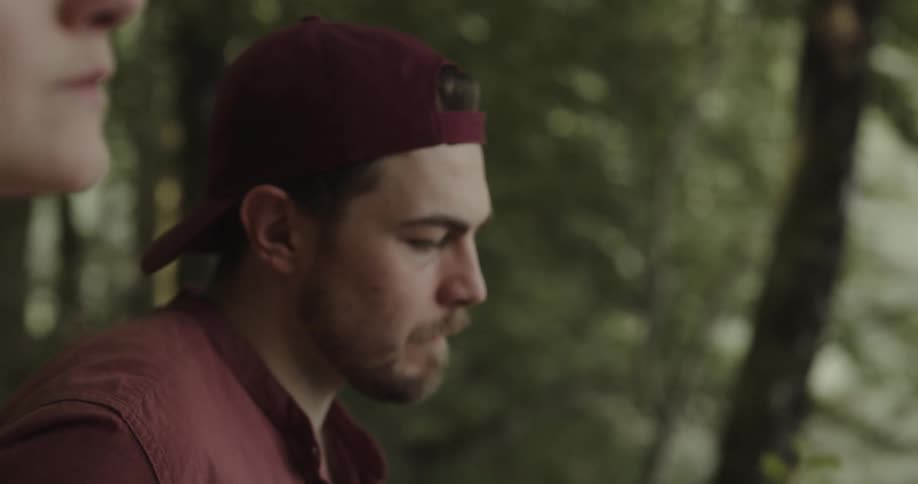 A woman is sitting alone in the woods and she looks upset and a man comes and sits near her