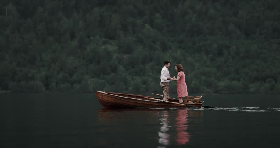Couple play in rowboat splashing and standing to bounce around near forest covered shore