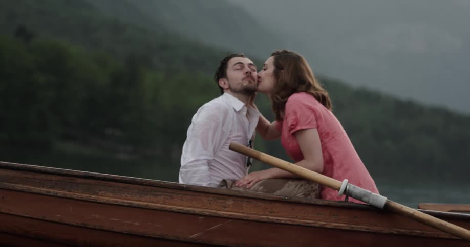 A man and woman in a rowboat touch their lips and cheeks as they ask one another for a kiss and then they laugh and kiss