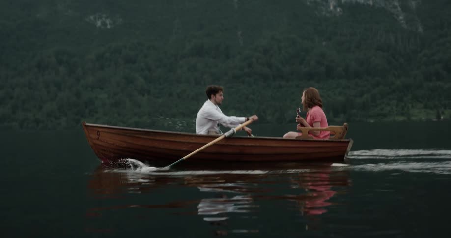 Man and woman in a wooden canoe in the middle of the lake while hes rowing and shes playing a mini guitar
