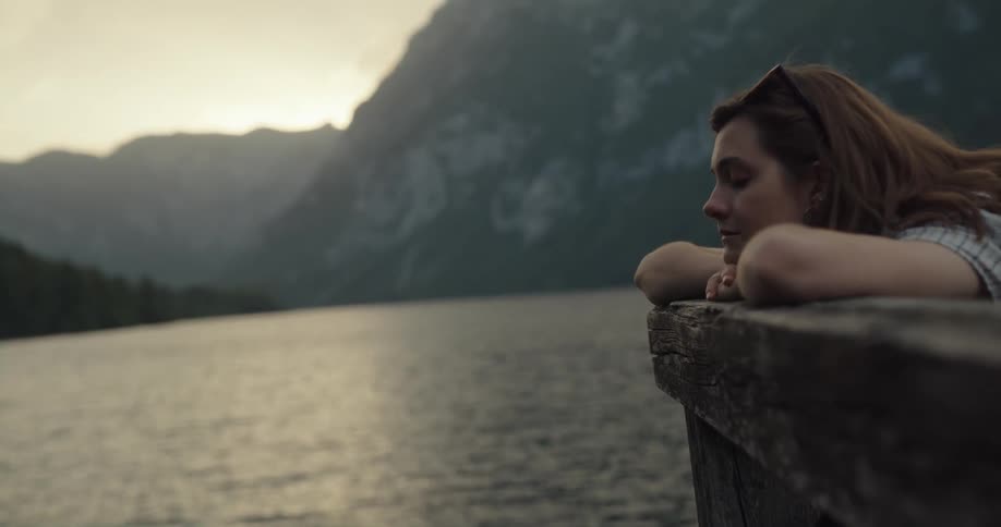Young woman reclined on the wood fence on a pier looking at a lake in the mountains at sunset