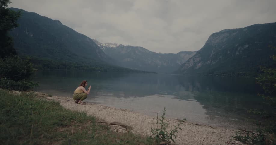 A woman kneels on the shore of a lake and raises her camera to take a photograph