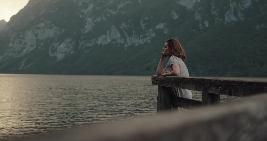 Young woman reclined on a wooden fence looks at a lake in the mountains at morning