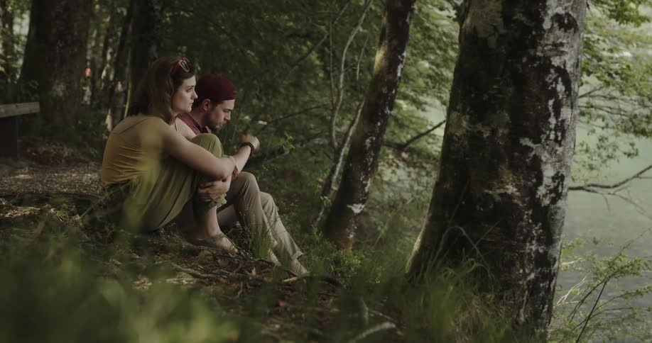A woman is sitting alone in the woods on a hill near a stream and she looks upset and a man comes and sits near her