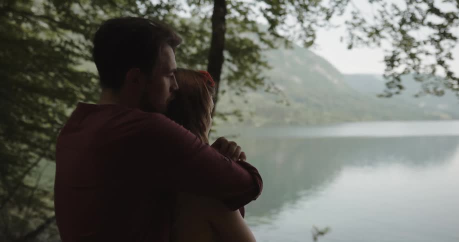 Man wraps his arms around a woman as they stand in a forest next to the lake