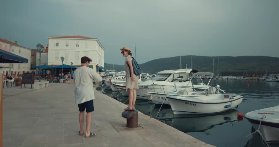 Boyfriend wearing white shirt and black shorts holds womans hands wearing hat sunglasses beige dress and blue backpack walking along the promenade and photographing her with camera