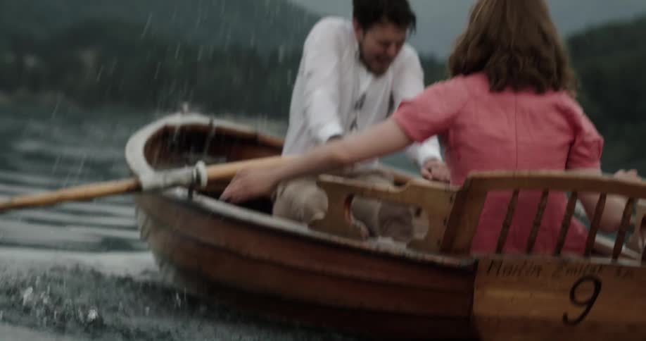 A woman riding in a boat on a lake with a man splashes the man with water as he rows the oars