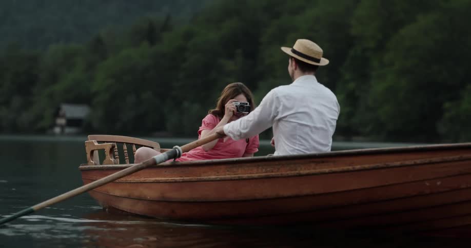 Man rows boat while woman takes pictures with forest and mountain in the background