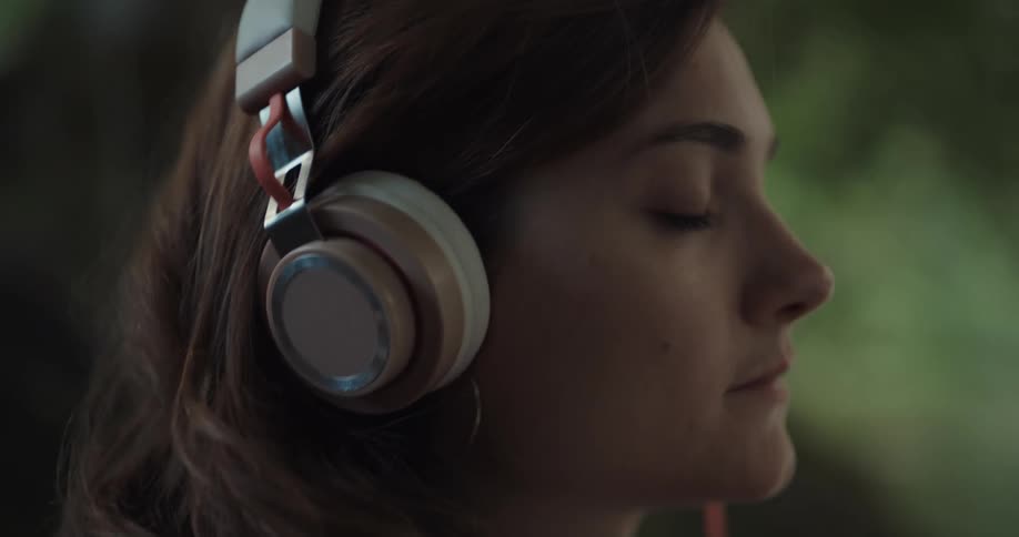 A woman listens to headphones with her eyes closed while sitting on a lakeshore during the day