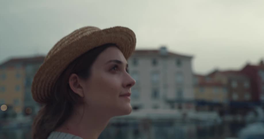 Woman with hat and dress walks along promenade and looks around at buildings in the town at dusk