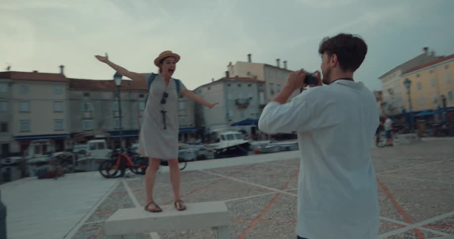 A woman poses atop a cement bench in a marina while a man photographs her with a camera