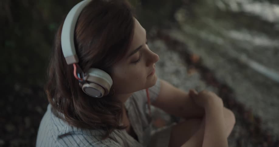 Young woman listening to music on headphones sitting on the shore of a lake at morning