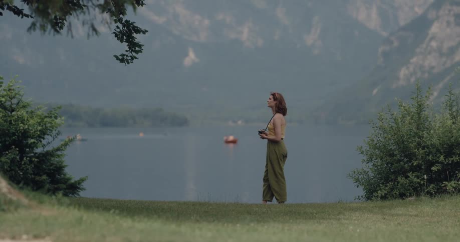 Woman standing next to a lake surrounded by mountains taking pictures of the place with a camera