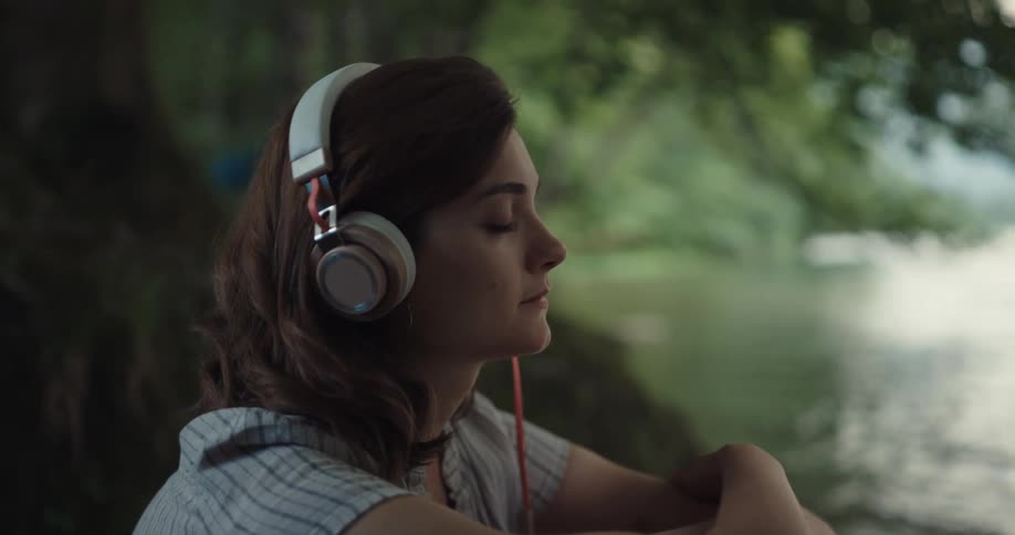 Woman sitting next to the water in a lake with her eyes closed listening to some music on her headphone