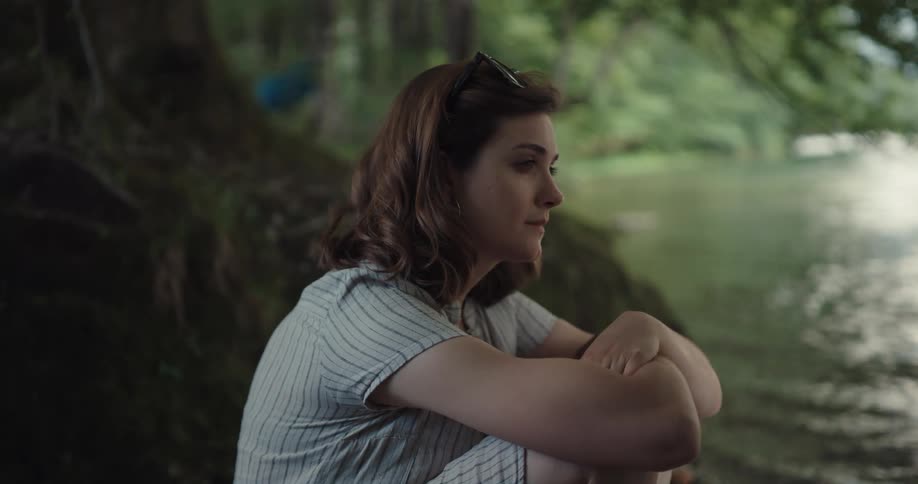 A young woman sits on the shore of a lake while lost in thought