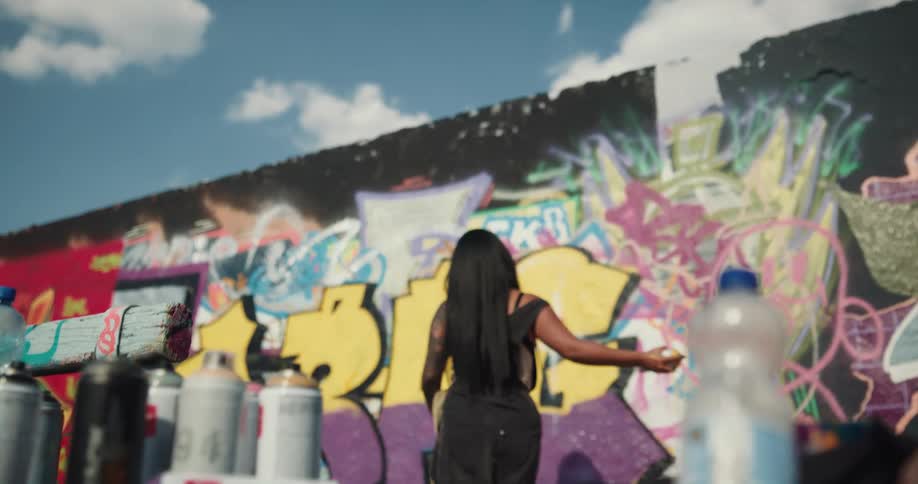 Black woman wearing a face mask grabbing a spray paint can and doing graffiti on the wall outdoors