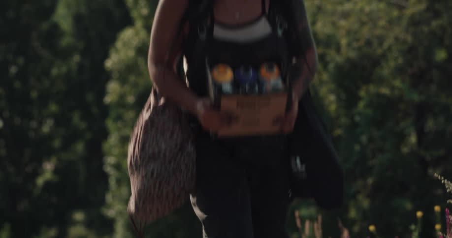 Black woman carrying a box filled with spray paint cans walking up the street in a sunny day