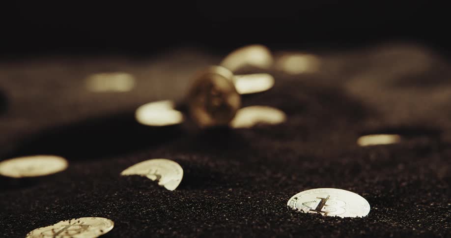 A number of shiny coins with the bitcoin logo on them are partially buried in black powder