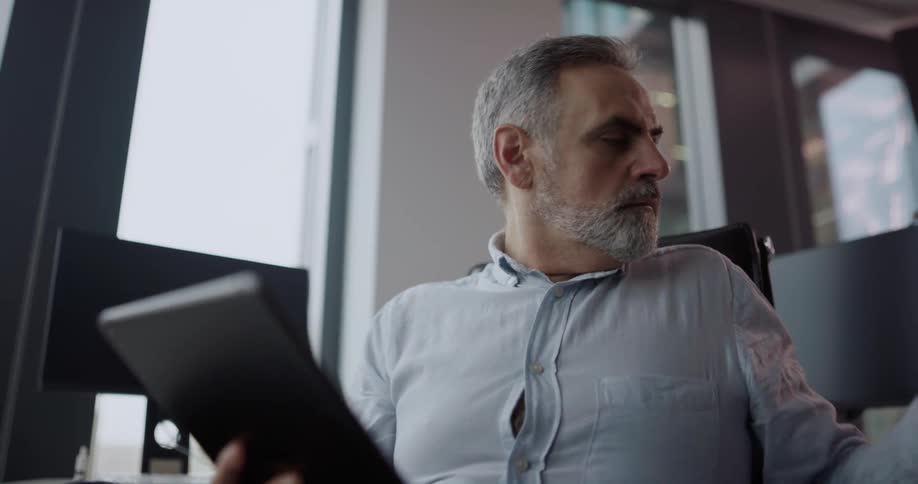 A man takes a sip from a coffee cup while looking at the screen of a computer tablet in a office during the day