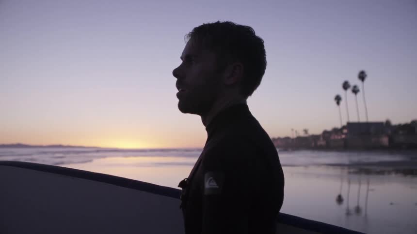 A man holding a surfboard is standing on the beach at sunset