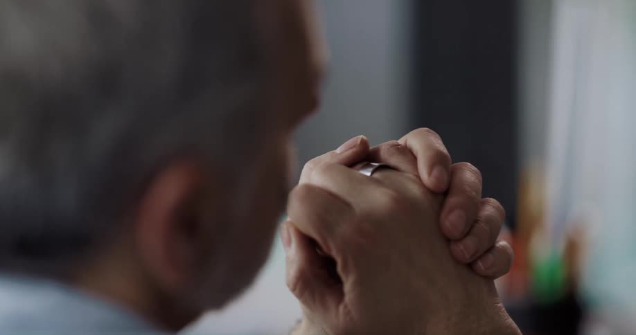 Old white man with grey hair and beard holding his hands together while listen to someone talking at an office space