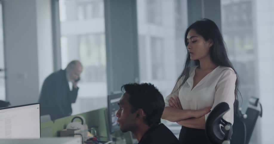 Man sits in a chair and looks at a monitor while a woman in a white blouse stands next to him and talks to him