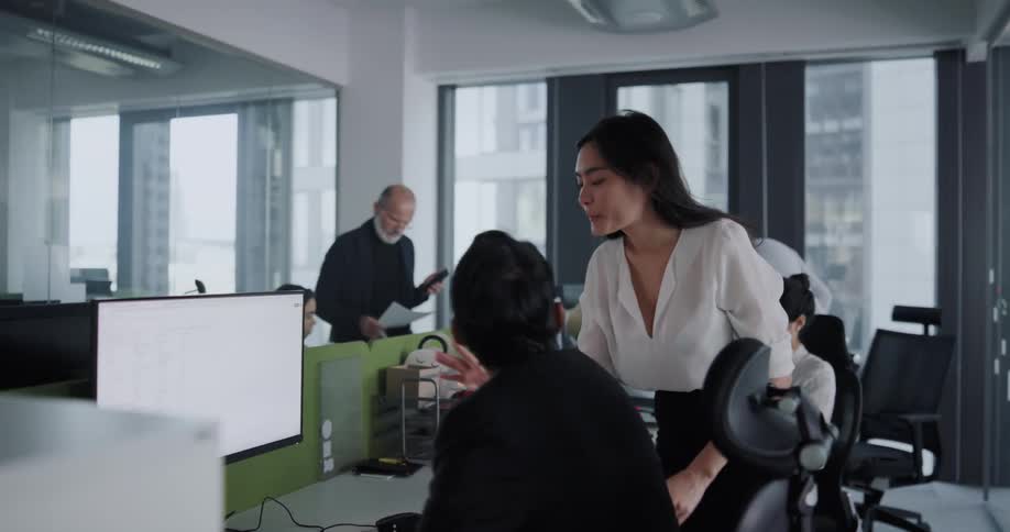 A woman watches a man using a computer at a desk in a shared office before leaning in to speak to him