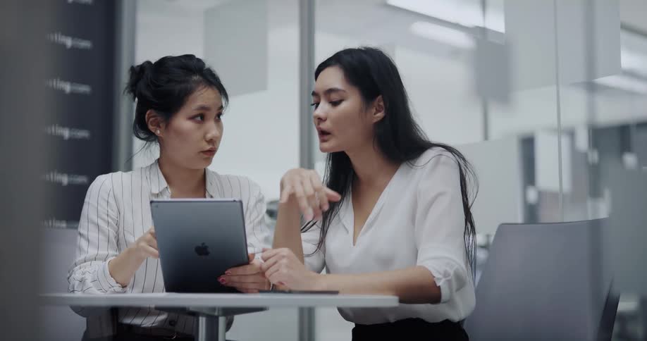 Two asian women sitting on a round table at a office space talking while using an ipad