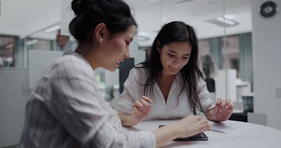 Two excited female coworkers sitting at a round table in an office space having a happy conversation while using a tablet