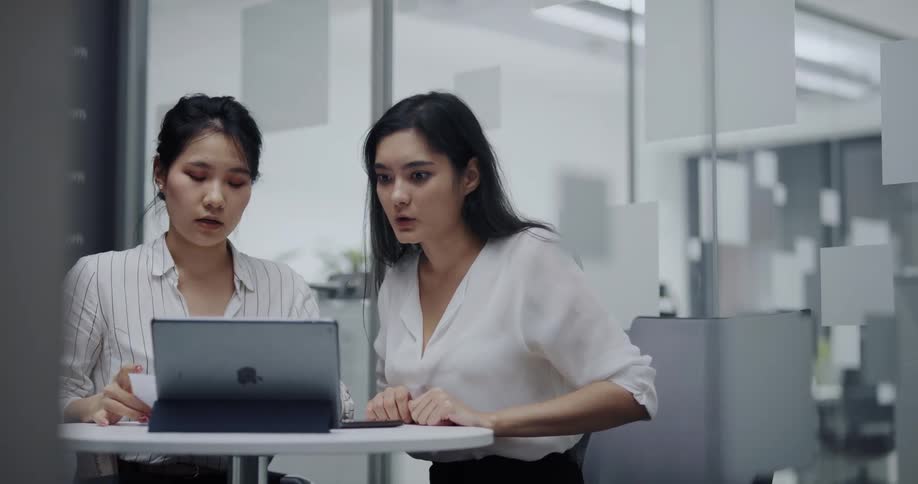 Two female coworkers sitting at a round table in an office having a video chat on an ipad