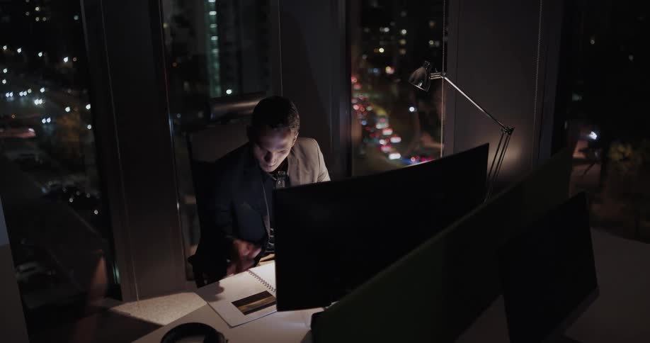 Businessman with black blazer jacket and shirt sits at desk works late hours in the office at night
