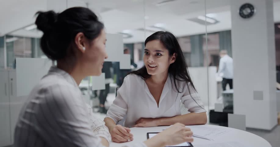 Two women sit at work and talk and look at a tablet and then high five one another