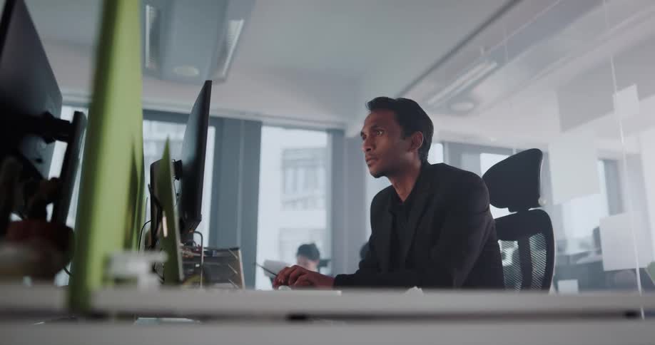 A man is in an office sitting at a desk watching something on the computer monitor and he appears to be pleased