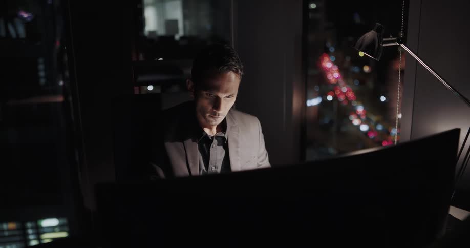 Ored businessman with black blazer jacket sits at desk and works late looking out office window at night