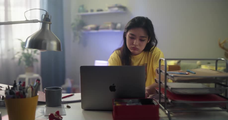 Teenage asian girl looking very tired sitting on her desk to study on her laptop and notebook