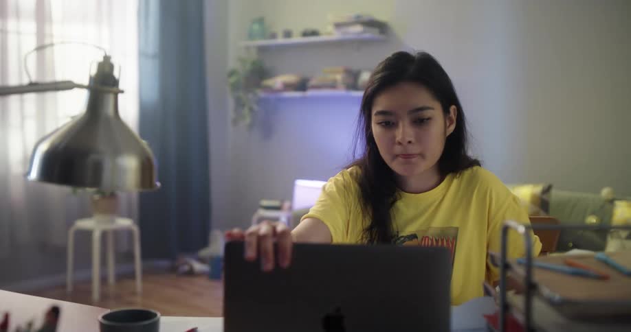 A young woman writing in a notebook while reading from a laptop closes her laptop and rubs her face before rising from her seat