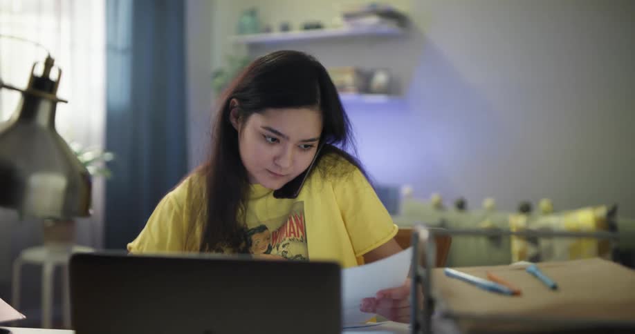 Young woman wearing yellow shirt working from home talks on the phone while checking paperwork