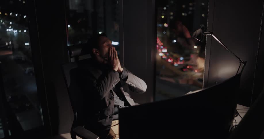 Bored businessman with black blazer jacket sits at desk and works late looking out office window at night