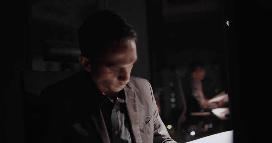 Office worker sitting at his desk next to a big glass window during the night looking very tired