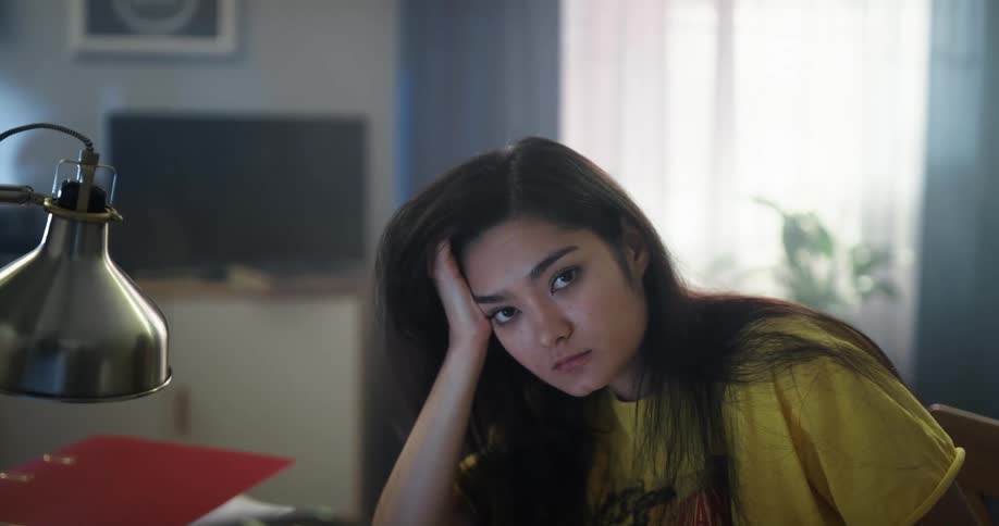 Bored teenage asian girl sitting at the desk resting her head on her hand