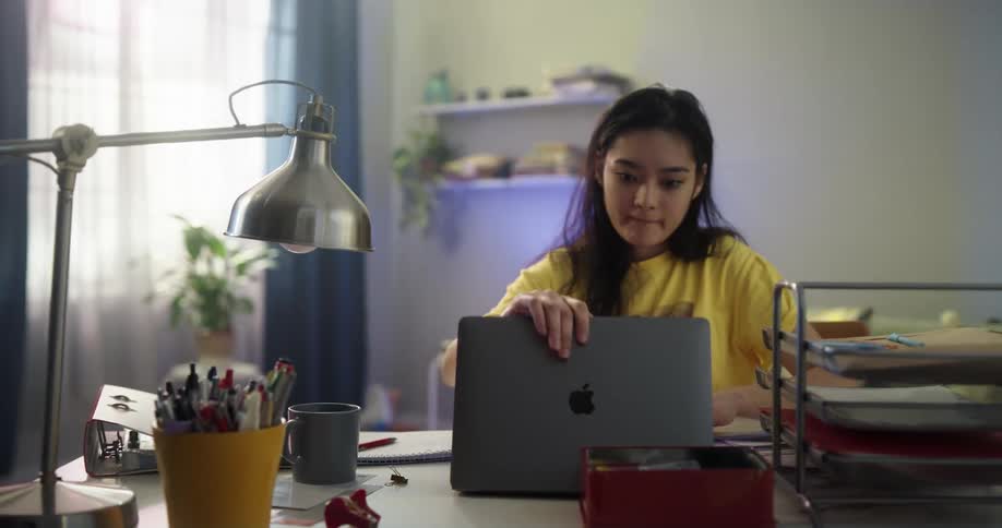 Teenage asian girl drinking a cup of coffee and then sitting on her desk opening up the laptop lid and start studying and writing down on papers