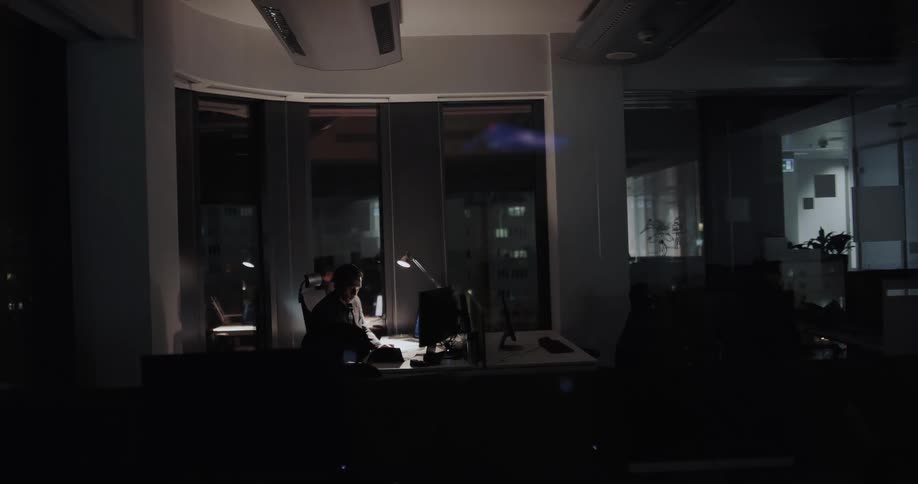 Businessman sitting at desk works on desktop and papers in office at night