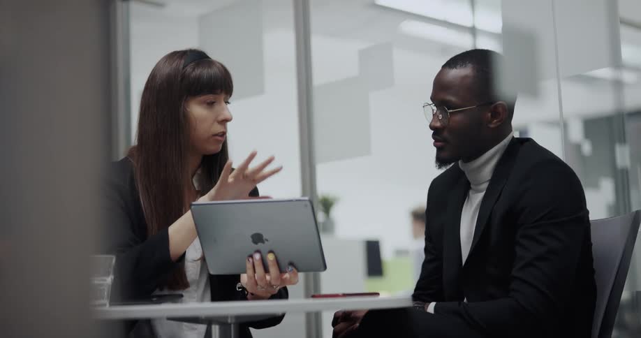 Woman holding an ipad while sitting at the desk in an office room talking to a man sitting on her side
