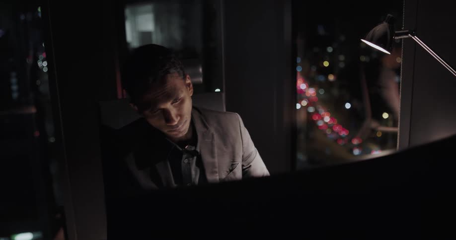 Bored businessman with black blazer jacket sits at desk and works late looking out office window at night